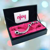 A curved, stainless steel Njoy 'Fun Wand' Kegel Exerciser & P-Spot Stimulator with three graduated bulbs is displayed in an open black box lined with bright pink fabric. The box has the word "Njoy" printed on the inside of the lid. The background is blurred with soft, colorful lights, perfectly highlighting this versatile product.
